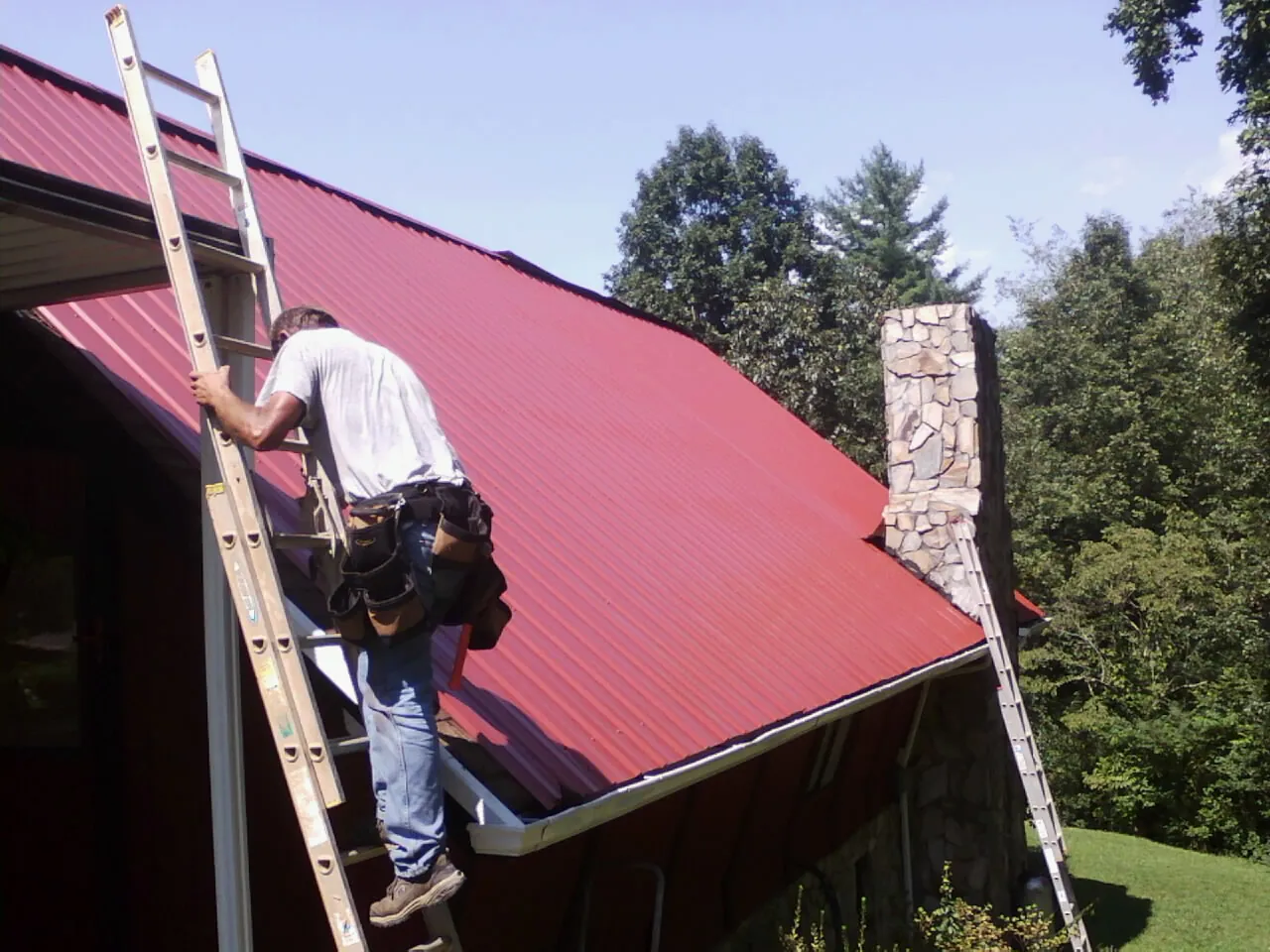 Roofer on a ladder beside a red metal roof and stone chimney — Soffit & Fascia Repair by Roofing Brothers in Matlock